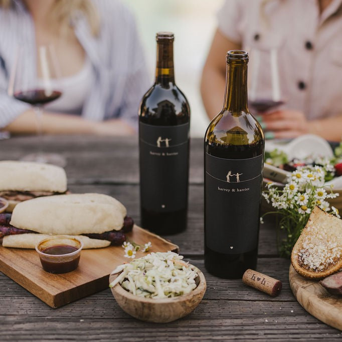 Two open bottles of 2021 Harvey & Harriet Red Blend on a rustic table with plates of food surrounding it.