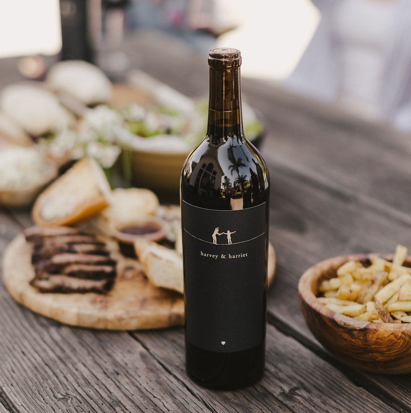 A bottle of 2021 Harvey & Harriet Red Blend on a rustic table with plates of food surrounding it.