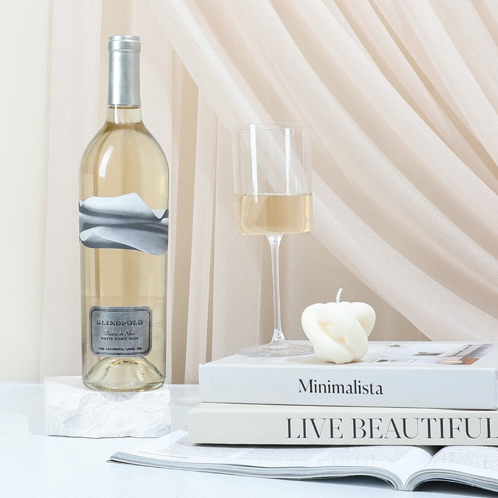 A bottle of Blindfold Blanc de Noir, books, and candle on a table with a soft curtain background