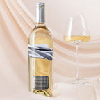 A bottle of Blindhold Sauvignon Blanc white wine stands next to a stemmed wine glass partially filled with the pale golden wine, set against a soft draped beige fabric background.