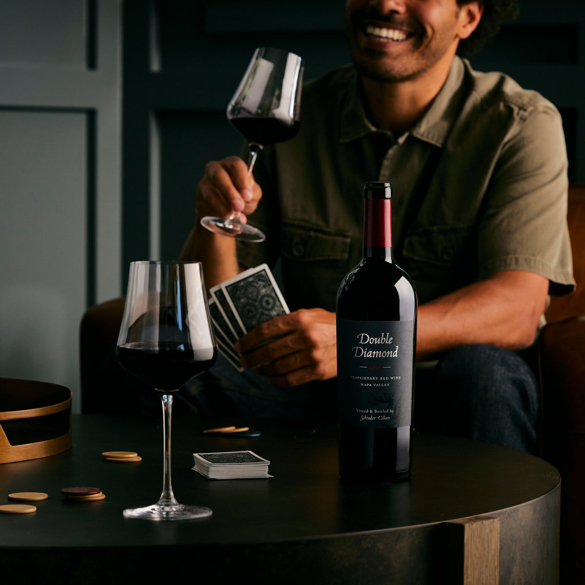 A bottle of Double Diamond : 2021 Napa Valley Proprietary Red Wine 750mL on a low table with a man playing cards and holding a glass of wine while smiling broadly