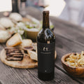 A dark glass bottle of Harvey & Harriet red wine stands on a rustic wooden table, surrounded by food like bread, fries, and grilled meat in a blurred outdoor setting.