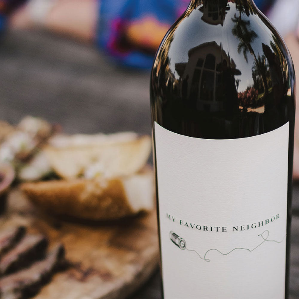 A close-up of a dark wine bottle with a white label reading 'MY FAVORITE NEIGHBOR' and a small line drawing, photographed outdoors with a blurred board of food and table in the background.
