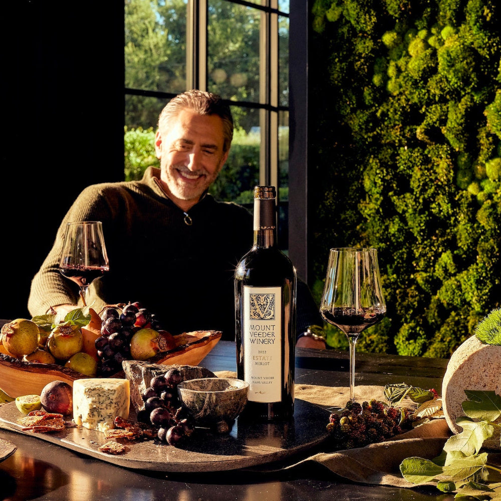 Man sitting at a table with wine and fruits, surrounded by greenery a bottle of Mount Veeder Estate Merlot in the foreground. 