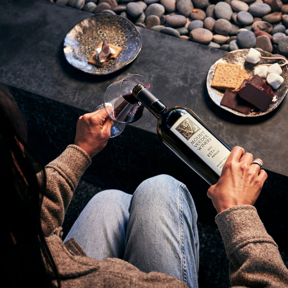 Person sitting at an outside with a bottle of Mount Veeder Winery Estate Merlot and a glass, surrounded by s'mores.