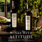 Two bottles of Mount Veeder Winery red wine, alongside a glass of red wine, set on a table with a green mossy background. WINES WITH ALTITUDE is printed at the bottom of the image