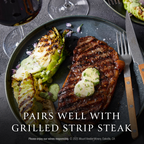 A plated grilled strip steak with butter on a dark plate, served with grilled cabbage and a fork, with wine glasses and a knife in the background. Text overlay reads 'PAIRS WELL WITH GRILLED STRIP STEAK'