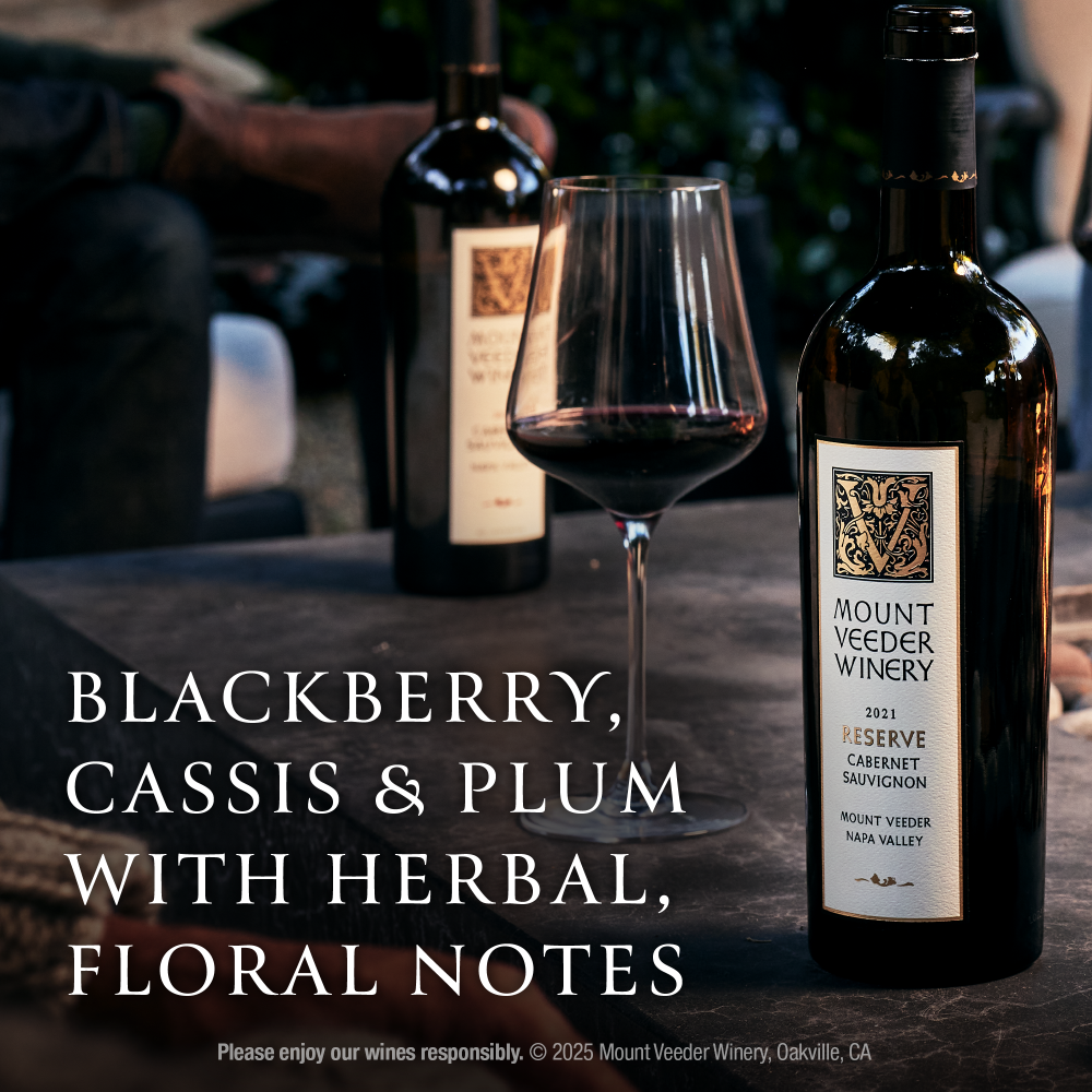 A bottle of Mount Veeder Winery 2021 Reserve Cabernet Sauvignon red wine stands on a dark table beside a wine glass and another blurred bottle in the background. Large text overlays describe the wine's flavor profile: blackberry, cassis, plum, and herbal, floral notes.