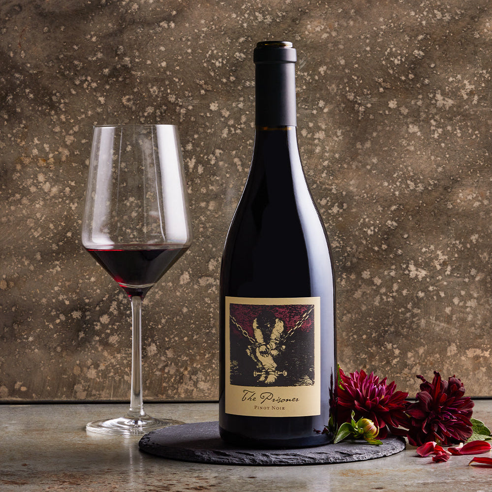 A bottle of the Prisoner Pinot Noir next to a wine glass with a textured background and some flowers
