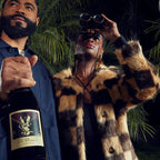 A man and woman stand in front of palm trees. The man holds a bottle of The Prisoner Pinot Noir, the woman is looking through binoculars