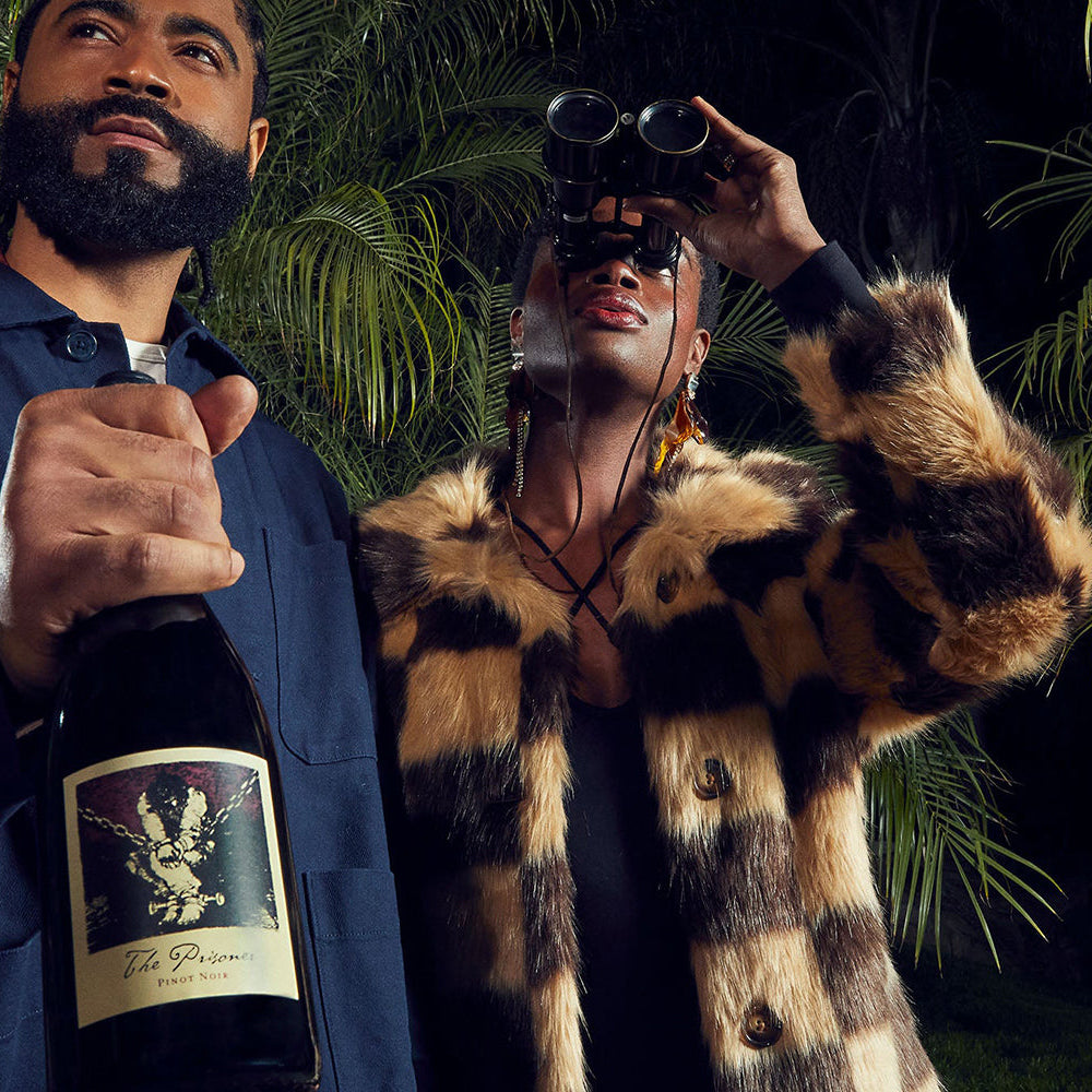 A man and woman stand in front of palm trees. The man holds a bottle of The Prisoner Pinot Noir, the woman is looking through binoculars