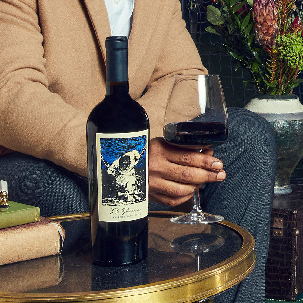 Person holding a wine glass next to a bottle of The Prisoner Cabernet Sauvignon and a decorative background