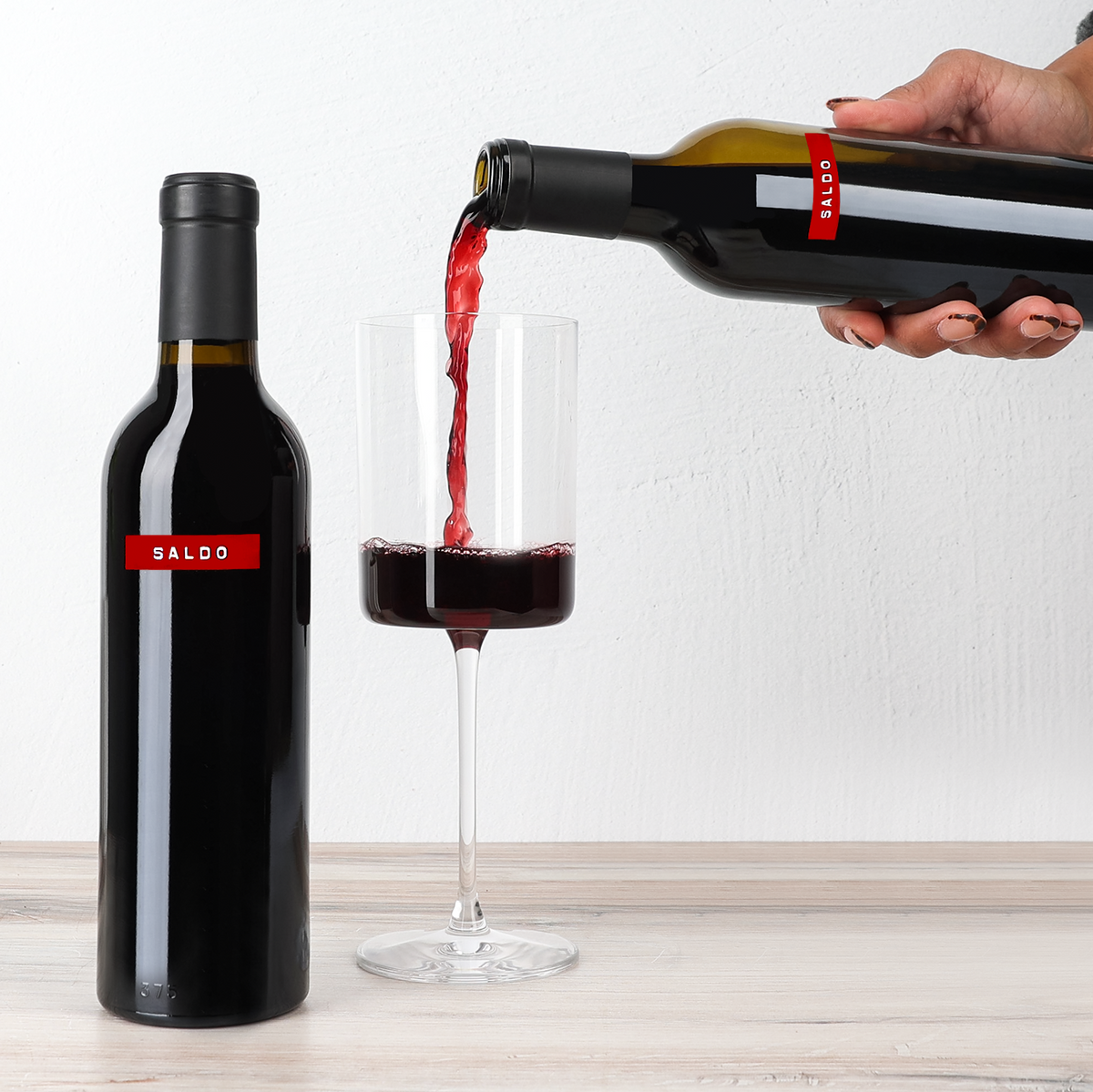 A hand pours red wine from a dark bottle labeled SALDO into a clear wine glass on a light wood surface against a white wall.