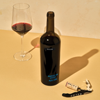 Bottle of Saldo Bavarian Lion Vineyard Petite Sirah on a yellow table with a wine glass and the cork on a corkscrew