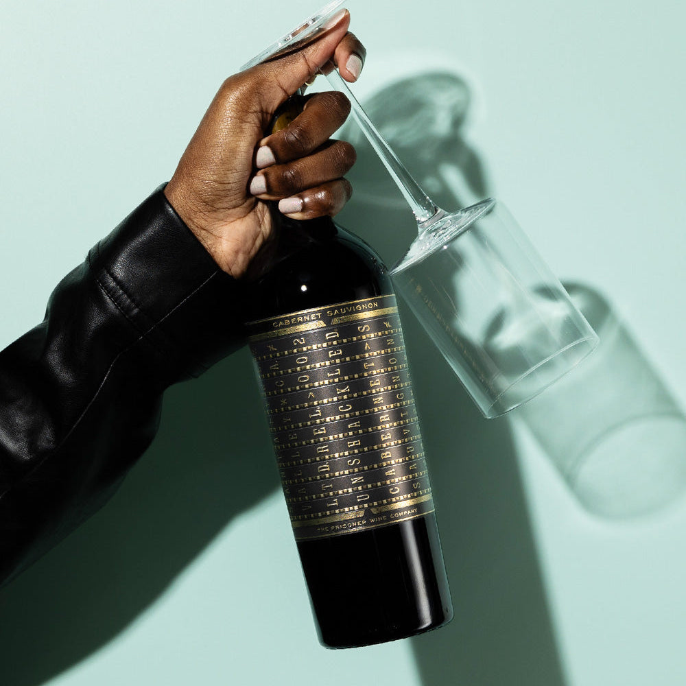 A woman holding a bottle of Unshackled Cabernet Sauvignon and a wine class in her had against a light green background