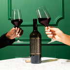 Two people holding wine glasses in front of a green door, with a bottle of Unshackled Cabernet Sauvignon between them.