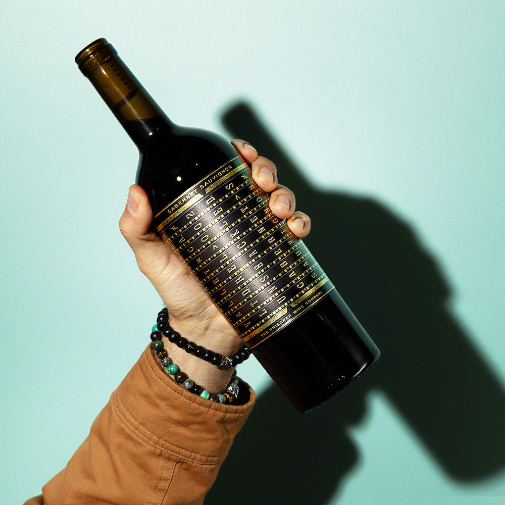 A mans hand holding a bottle of Unshackled Cabernet Sauvignon against a light green background with a strong shadow.