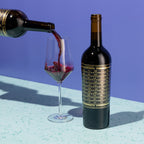 Red wine being poured from a bottle into a glass with a bottle of Unshackled Cabernet Sauvignon on blue background 