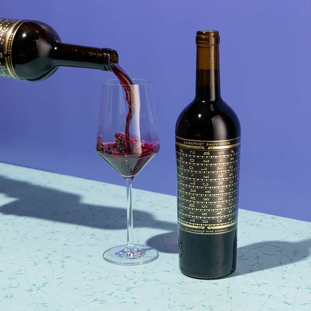 Red wine being poured from a bottle into a glass with a bottle of Unshackled Cabernet Sauvignon on blue background 