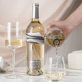 Bottle of white wine being poured into a glass with a bottle of Blindfold Blanc de Noir in the blurred background