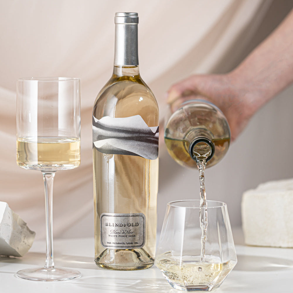 Bottle of white wine being poured into a glass with a bottle of Blindfold Blanc de Noir in the blurred background
